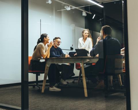 Four office workers in casual attire talk around a table in a collaborative workspace