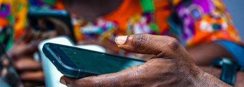 A man holding a cell phone. Behind him, someone in bright clothing.